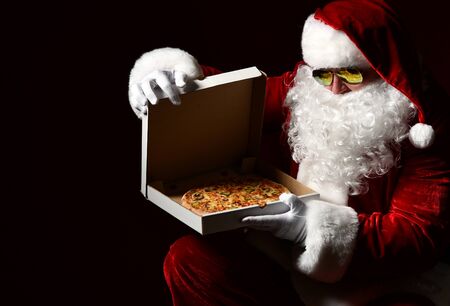 Santa Claus is opening closing pizza box, looking inside, desiring, going to take a slice, packing gift on black with copy space. New year and Merry Christmas fast food conceptの写真素材