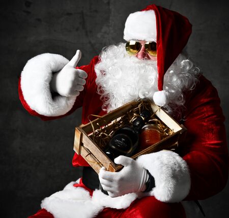 Santa Claus in sunglasses is holding a crate with a canned red salmon caviar and luxury champagne wine bottle showing thumb up sign on dark. New year and Merry Christmas conceptの写真素材