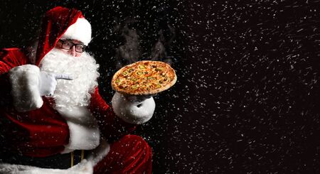 Happy laughing Santa Claus hold big hot steaming original pizza offering pointing finger under the snow with copy space. New year and Merry Christmas fast food concept bannerの写真素材