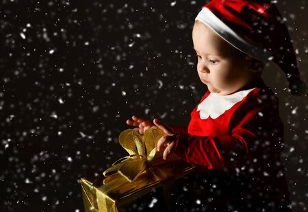 Infant baby boy toddler is sitting in red christmas cap and with xmas gift box with golden ribbon bow playing opening present under the snow on dark backgroundの写真素材