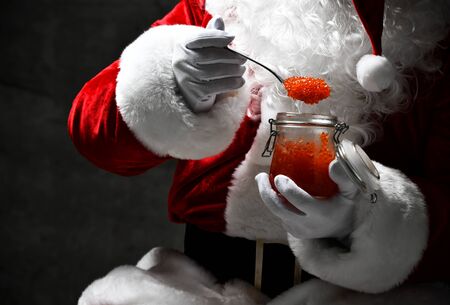 Closeup portrait of Santa Claus eating  red caviar fish salmon looking at spoonful, holding a jar on dark gray concrete wall background. Happy new year and Merry Christmas conceptの写真素材