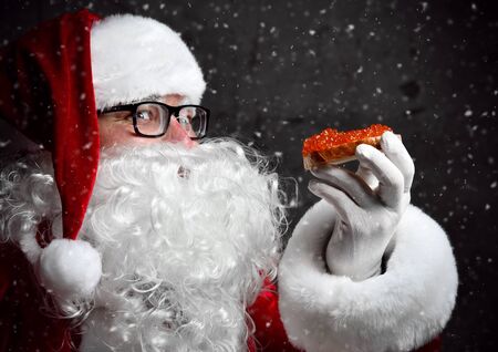 Santa Claus is holding red salmon caviar sandwich, looking glancing at us, going to eat under the snow on dark background. New year and Merry Christmas and happy holidays conceptの写真素材