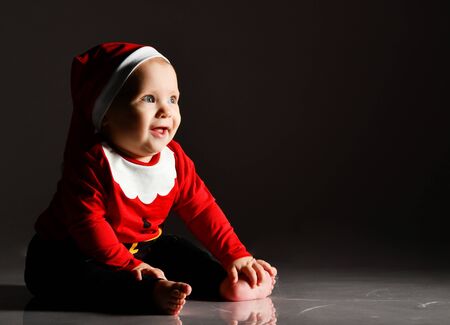 Smiling infant baby boy barefooted toddler in red christmas cap and new year costume is sitting on...