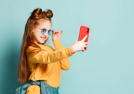 A beautiful red-haired girl takes a selfie with a new mobile phone in a red case. In blue glasses and a yellow sweater with a hood on a blue backgroundの写真素材