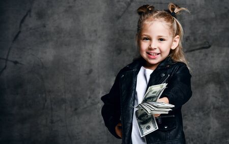 Happy screaming rich kid girl lucky millionaire in leather jacket is holding a bundle of moneyの写真素材