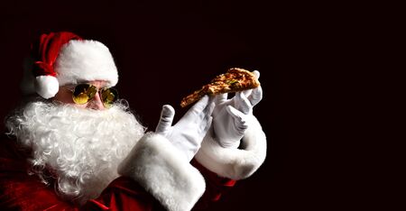 Santa Claus in aviator sunglasses is holding tasty slice of pizza, looking at it, going to eat on darkの写真素材