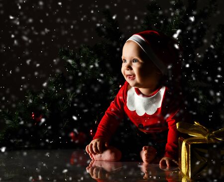 Infant  boy  in red christmas cap and costume is sitting at golden New Year gift box present with ribbon and bowの写真素材