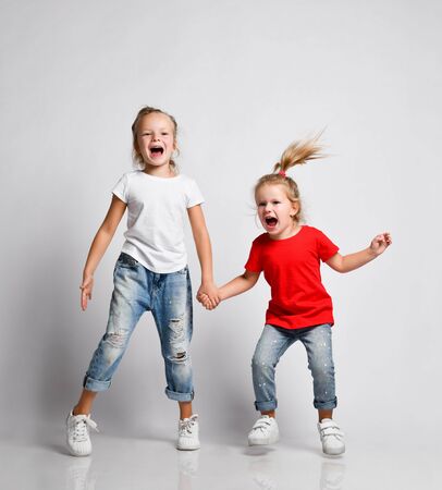 Two kids blonde girls sisters friends in jeans and t-shirts are jumping hand in hand, singing, relieving.の写真素材