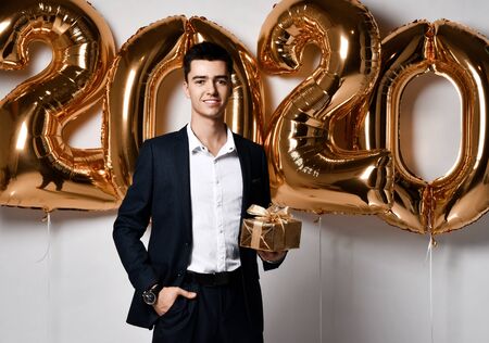 Young guy businessman in an expensive suit holds a Christmas gift happy on a gold foil balloons...
