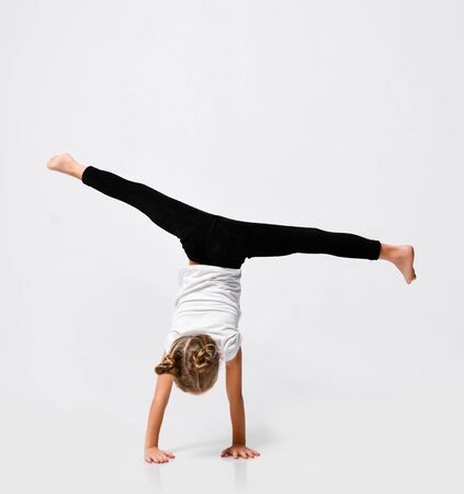 Slim athletic blonde kid girl is doing gymnastic acrobatics exercises, handstand and stretching with her legs up on white with free copy spaceの写真素材