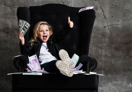 Happy lucky rich kid girl in leather jacket is sitting in luxury armchair with a bundle of money dollars cash showing thumb up sign gesture, screaming on concrete wall backgroundの写真素材