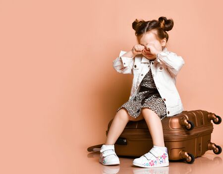 Offended kid girl in leopard print dress, denim jacket has got lost. She is sitting on roller suitcase bag on wheels and cries wiping away a tear on background with free copy spaceの写真素材