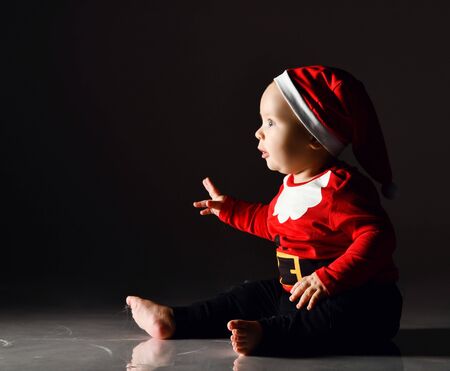 Amazed infant child baby boy toddler in red santa claus costume and cap is sitting sideways to us holding his handの写真素材