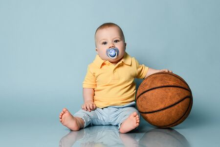 Cool infant baby boy with pacifier and in jeans and yellow shirt is sitting on the floor holding basket ball on background with free copy spaceの写真素材
