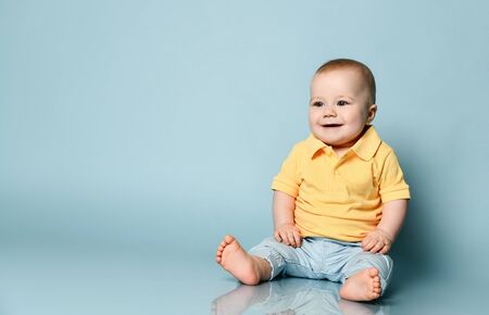 Happy smiling barefooted Infant baby boy toddler in yellow shirt and blue jeans is sitting on the floor looking forward on background with free copy spaceの写真素材