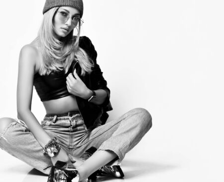 Portrait of young grunge blonde woman in torn jeans, top, brutal boots and winter hat sitting cross legged, holding leather jacket on floor on white background with copy spaceの写真素材
