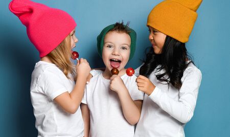 The company of three cheerful friends boy and two girls in colorful hats standing close together, looking at each other and eating sweet lollipops on blue mint backgroundの写真素材