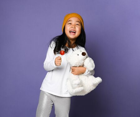 Happy asian kid girl playing with polar bear toy and eating lollipop candy happy smiling screaming jumping on one leg on purple. International day of polar bear concept.の写真素材