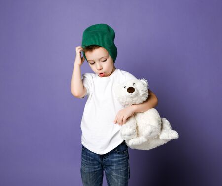 Young concerned kid boy child in green hat holding hugging his best friend polar bear toy looks scratches his head. International day of polar bear concept on purple backgroundの写真素材