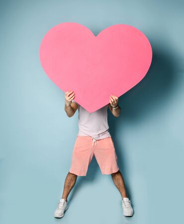 Young man hide his face behind pink heart shape toy in hands on pastel blue wall background. Love gift for valentines day with text spaceの写真素材