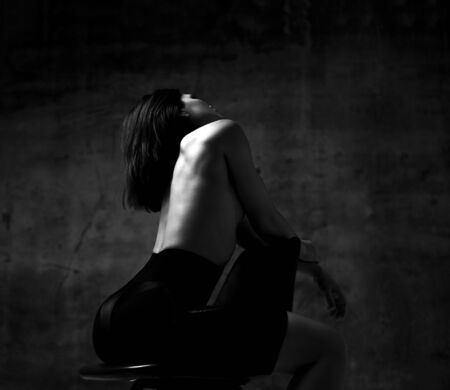 Young slim beautiful brunette woman in black sport shorts sitting backwards on chair with naked back over dark wall background. Stylish sexy sporty outfit and beauty of womans body conceptの写真素材
