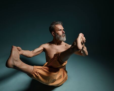 Old man with grey beard doing yoga, pilates, fitness training, stretching exercise, asana or balance workout on floor over blue background. Healthy lifestyle conceptの写真素材