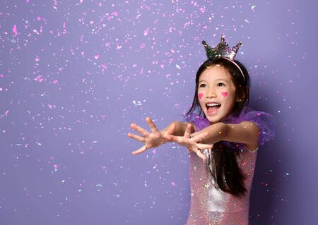 Happy laughing asian kid girl princess in party dress and with a painted red hearts sign on cheeks is stretching out her hands touching snowflakes on purple backgroundの写真素材