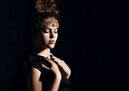 Attractive brunette woman with golden tiara crown on her forehead beam hairstyle in black smart body, nylon tights posing sexy on dark wall backgroundの写真素材