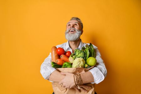 Cool old mature senior man with gray beard shopping hold grocery shopping bag with healthy organic vegetables looking up on yellow backgroundの写真素材