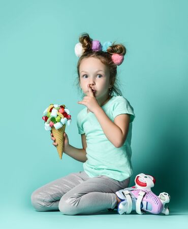 Little kid girl in roller skates sits on floor holding ice cream in waffles cone with sweets marshmallows candies holding finger at her lips makes shhh signの写真素材