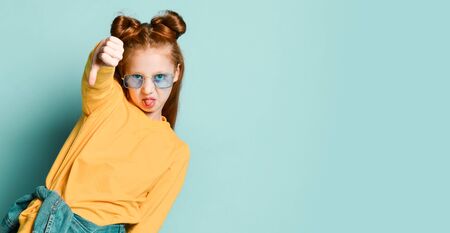 Red-haired teenager girl in smoked blue glasses and yellow sweatshirt is showing thumbs down sign gesture on background with free text copy spaceの写真素材