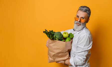 Cool old mature senior man with gray beard shopping hold grocery shopping bag with healthy organic vegetables on pastel pink backgroundの写真素材