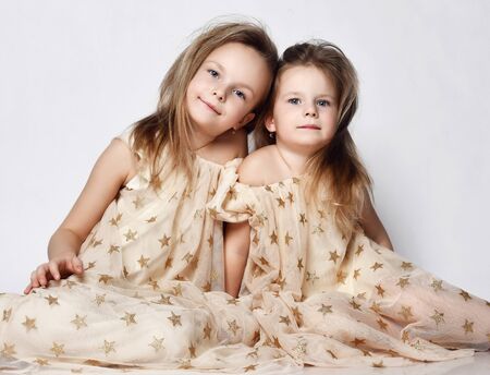 Two pretty little sisters hugging and sitting in beautiful dresses with stars. Toddler kids with blue eyes. Cute girls relax on white background. Love, trust and tenderness.の写真素材