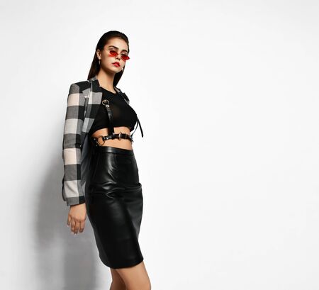 Young brutal brunette woman in checkered jacket, black leather skirt, cross-belt and narrow sunglasses is sexy posing over white background with free copy spaceの写真素材