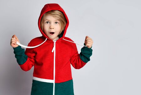 Portrait of frolic excited surprised blond kid boy in dark gray and red hoodie with his cowl over his head on gray background with free copy spaceの写真素材