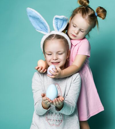 Two small happy girls in cosy home clothing and decorative fur ears standing, hugging and holding white and beige eggs in hands over blue wall background. Family look and celebrating Easter conceptの写真素材