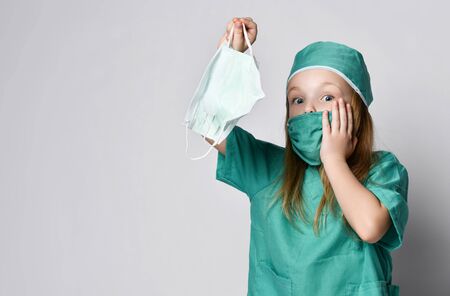 Little girl child play doctor in green medical coat hold  protection masks from the Coronavirus Covid-19 surprised. Madness pandemic panic epidemic spreads across the globeの写真素材
