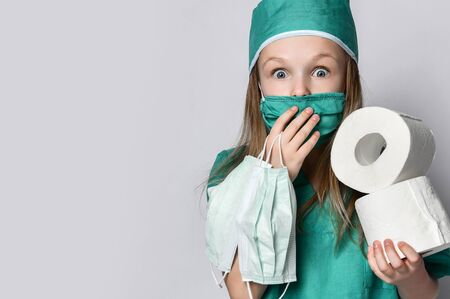 Little girl in a medical coat holds bactericidal protection mask from the Corona Virus and toilet paper rolls surprised. Madness pandemic Coronavirus Covid-19 panic epidemic spreads across the globeの写真素材