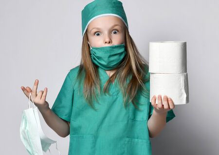 Little girl in a medical coat holds bactericidal protection mask from the Corona Virus and toilet paper rolls surprised. Madness pandemic Coronavirus Covid-19 panic epidemic spreads across the globeの写真素材