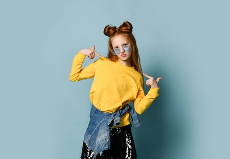 Stylish red-haired teen girl in yellow sweatshirt and glossy black skirt is posing as a top model pointing fingers at herself with both hands saying I am the best and coolの写真素材