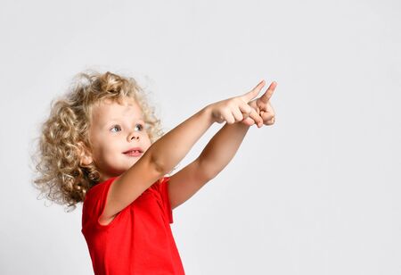 Portrait of curly hair blonde kid girl in red t-shirt holds pointing fingers with both hands at free copy space at upper cornerの写真素材