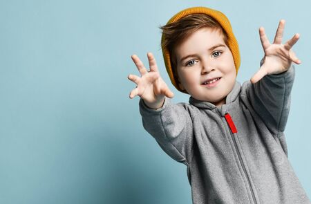 Little brunet male in orange hat and gray overall. He is smiling, reaching his hands to you, posing against blue studio background. Childhood, fashion, advertising. Close up, copy spaceの写真素材