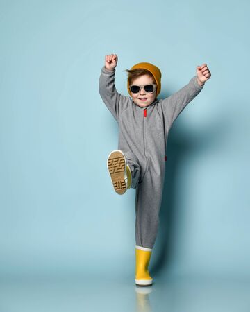 Brunet boy in sunglasses, orange hat, gray overall and yellow rubber boots. He smiling, raised hands and leg, posing on blue studio background. Childhood, fashion, advertising. Full length, copy spaceの写真素材
