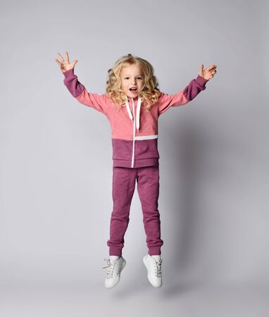 Playful frolic screaming kid girl in stylish modern pink gray sportswear hoodie and pants jumps high with her hands up over white background with free text copy spaceの写真素材