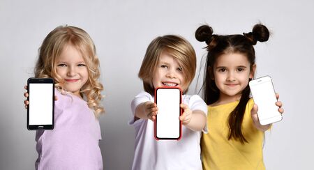 Two smiling little baby girls and boy in stylish casual clothing standing and showing smartphones screens over grey wall background. Happy childhood and technologies conceptの写真素材