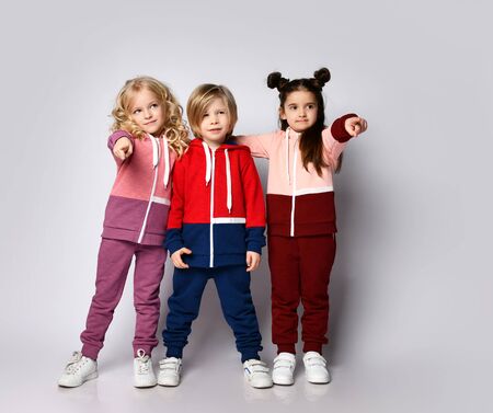 Full growth portrait of three kids blonde and brunette girls and blond boy in modern hoodies and pants stand together pointing fingers at usの写真素材