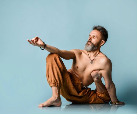 Old man with beard in trousers doing yoga, pilates, fitness training, stretching exercise, asana or balance workout over blue background, top view. Healthy lifestyle and body opportunities conceptの写真素材