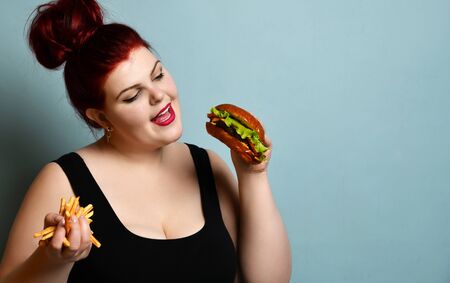 Happy overweight fat woman happy hold burger cheeseburger sandwich with beef and french fries in hand on pastel turquoise background. Healthy eating diet fast food conceptの写真素材