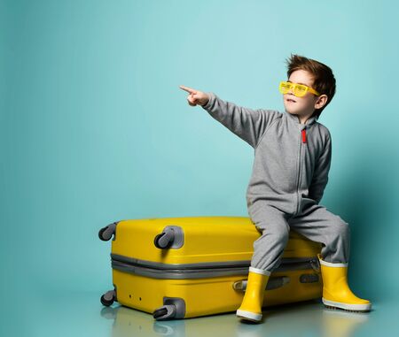 Little brunette kid in gray overall, yellow sunglasses and rubber boots. He sitting on suitcase, pointing at something, posing on blue background. Childhood, fashion, travelling. Full length, copy spaceの写真素材