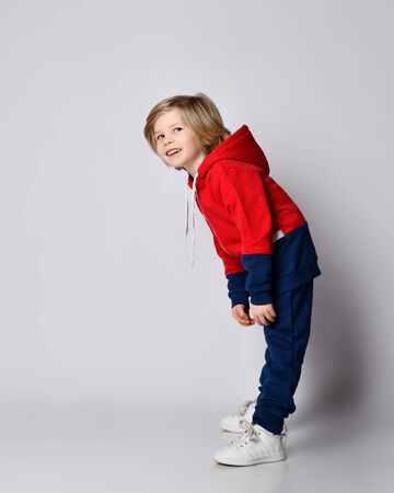 Full growth portrait of active blond kid boy in blue and red sportswear hoodie and pants doing exercises bending over reaching his hands to his toesの写真素材